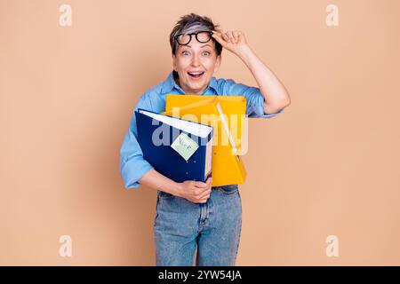 Charming senior woman holding colorful folders with a cheerful expression on beige background Stock Photo