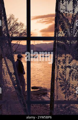 View through curtains of man overlooking boats on lake at sunset Stock Photo