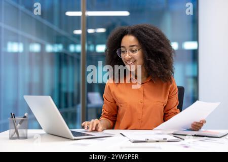Confident professional woman office, reviewing documents and using a laptop. Showcasing positivity and productivity in her work environment, creating a dynamic and inspiring office scene. Stock Photo