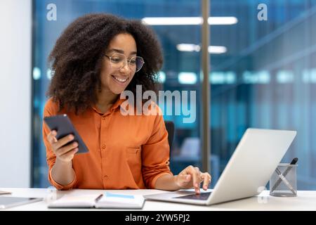 Young woman multitasking with a smartphone and laptop at her office desk, displaying a cheerful and productive work attitude. The image depicts a professional setting with modern technology in use. Stock Photo