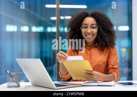 Smiling woman in an office setting, holding an envelope, sitting at a desk with a laptop and notebooks. The scene conveys professionalism, focus, and a positive working environment. Stock Photo