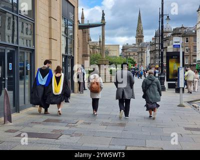 Two college students in graduation gowns (academic regalia) walking on sidewalk in Dundee, Scotland UK next to other people; seen from the back Stock Photo