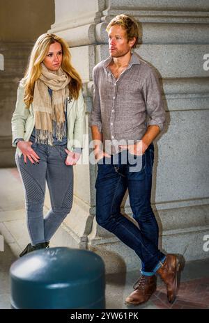 Two people are leaning casually against a stone column, both dressed in stylish, casual attire. The setting appears to be urban and well-lit, enhancin Stock Photo