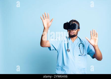 Nurse wearing virtual reality headset remotely operating patient. Healthcare specialist using high tech futuristic modern medical vr gadget, standing against blue background in studio. Stock Photo