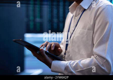 Engineer working in high tech server farm, using laptop and tablet to analyze data, ensuring seamless data flow. Man examining hardware in data center, ensuring system integrity and security, close up Stock Photo