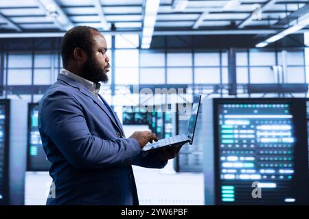 Programmer using laptop in data center with advanced technology, ensuring cybersecurity and efficient network management. Professional in server hub optimizing data center infrastructure with notebook Stock Photo