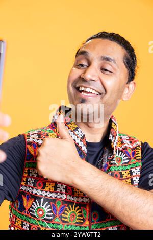 Happy man using cellphone to take selfies, doing thumbs up gesture. Jolly social media user taking photos using phone selfie camera, doing like hand gesturing, studio background Stock Photo