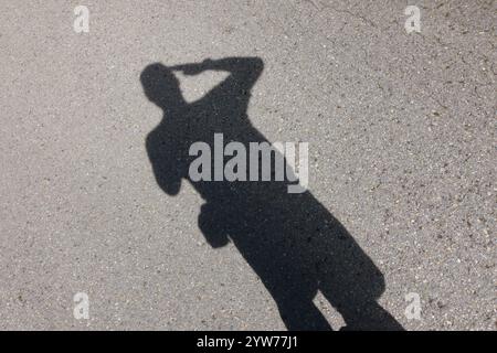 Human silhouette shadow cast on gray asphalt pavement, person in dynamic pose with arms raised overhead, sharp contrast under bright sunlight, artisti Stock Photo