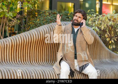 Indian young businessman tourist having bad stressful news calling on smartphone. Sad frustrated confused Lebanese bearded guy have problem unpleasant remote phone conversation on downtown city street Stock Photo