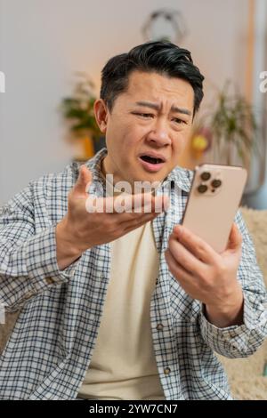 Upset displeased Chinese young man with brown hair using smartphone sitting on sofa in living room apartment. Middle-aged guy in casuals loses becoming surprised by sudden lottery results or bad news. Stock Photo
