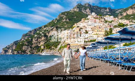 A diverse couple enjoys a romantic walk along the pebbled Amalfi Coast beach, surrounded by colorful houses on a hillside. The sky is clear and blue, perfect for an unforgettable day by the sea. Stock Photo