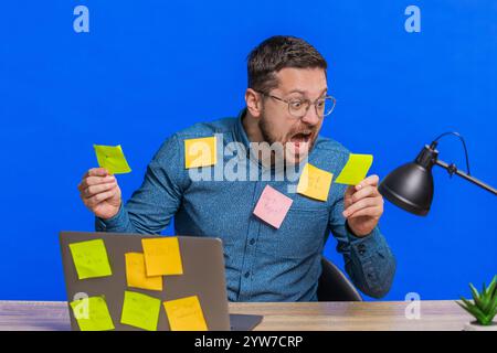 Exhausted man sitting at table with pasted sticker notes use laptop computer having concentration problem creative crisis. Nervous adult guy confused by big routine work loading on blue background Stock Photo