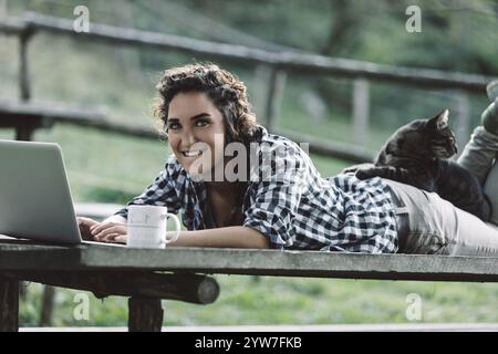 Young woman lying on a wooden picnic table, working on her laptop while sipping coffee. A cat comfortably resting on her back adds a cozy touch to the Stock Photo