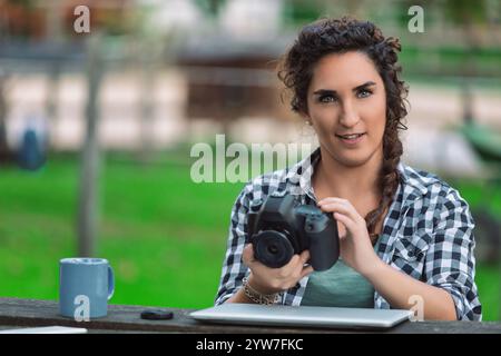 Young female photographer sitting at a table in a park, checking photos on her digital camera while enjoying a cup of coffee and working on her laptop Stock Photo