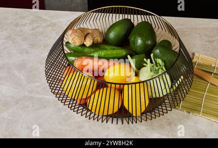 A decorative wire basket filled with vibrant vegetables and fruits, including avocados, lemons, carrots, and ginger, on a marble counter. Stock Photo