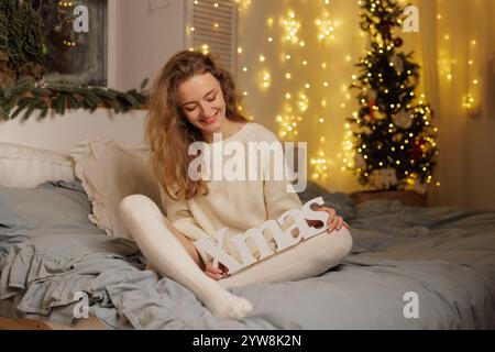 A young woman sitting on a bed, smiling while holding a decorative sign that says 'Xmas'. The room is decorated with fairy lights and a Christmas tree Stock Photo