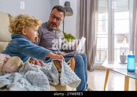 Father multitasking with work and family, using laptop while caring for toddler son in cozy living room Stock Photo