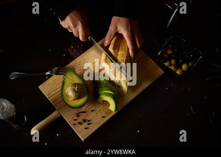 A person slices bread and avocado on a wooden board in a dimly lit setting, creating a rustic and minimalistic atmosphere. Stock Photo
