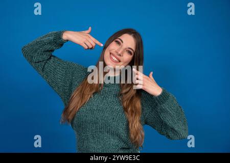 Studio portrait of cheerful young woman pointing fingers at herself, smiling on blue background Stock Photo