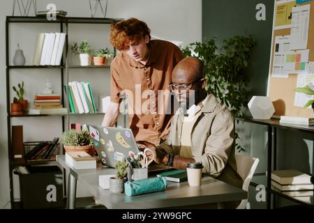 Red haired man watching his African American colleague explaining new codes on laptop in office Stock Photo