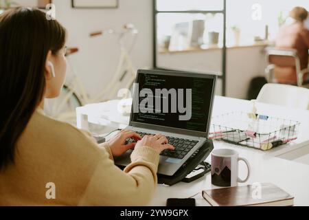 Back view of unrecognizable female office worker sitting at table and writing code on laptop with cooling pad Stock Photo