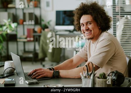 Medium close up male IT specialist smiling widely while looking at camera, he sitting at work desk with hands on keyboard Stock Photo