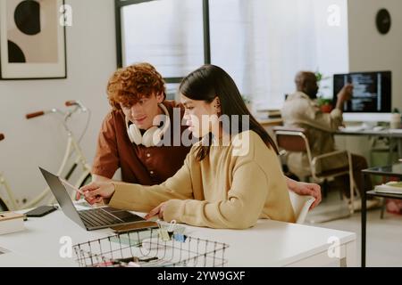 Female and male colleagues sitting at table and looking at monitor while young adult brunette woman showing presentation of brand new project Stock Photo