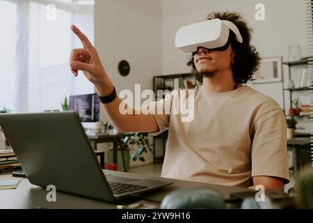 Low angle of young adult man sitting at desk with laptop and gesticulating while using VR goggles Stock Photo