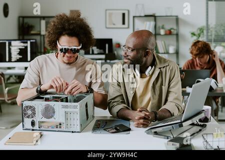 Biracial friends chatting while working in office, African American worker watching his biracial colleague connect wires of old computer Stock Photo