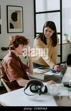 Vertical shot of young adult brunette female developer pointing at laptop screen while her coworker looking at it thoughtful Stock Photo