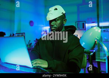 Medium close up of African American hacker wearing black hoodie and mask on forehead coding on laptop in dark room with neon lights behind him Stock Photo