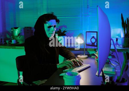 Curly haired man with white mask hiding his face while programming virus via computer in room lit by neon green and turquois lights Stock Photo