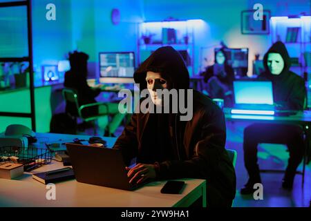 Medium close up of anonymous man in mask sitting at desk with laptop on top and coding in dimly lit room with digital devices Stock Photo