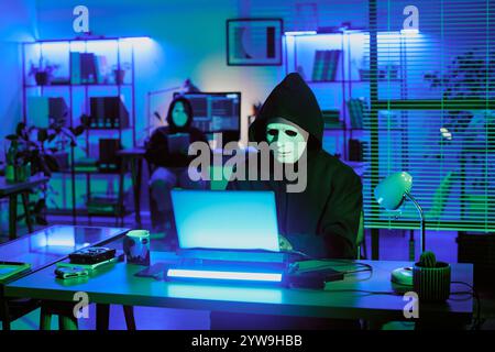 Medium close up of unrecognizable man with white mask programming via laptop in neon office Stock Photo