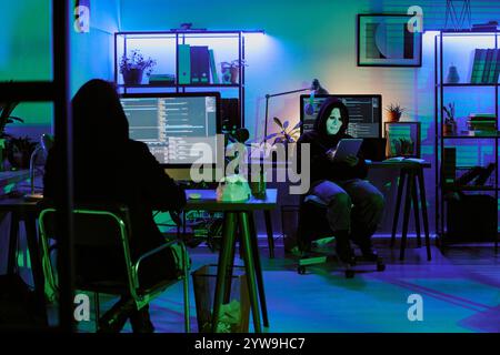 Unrecognizable people in jet black hoodies sitting at work desk and programming secretly Stock Photo