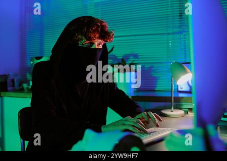 Medium close up ref curly haired man wearing black mask to hide his face sitting at table and hacking computer Stock Photo