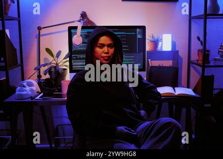 Portrait of female hacker dressed in black hoodie smiling lightly, table with computer, personal stuff and white mask behind her in dimly light room Stock Photo