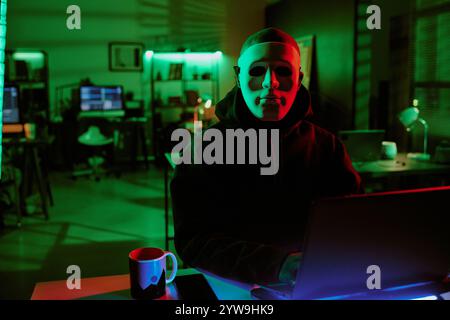 Medium close up portrait of unrecognizable man wearing white mask and sitting at desk with laptop and smartphone on top Stock Photo