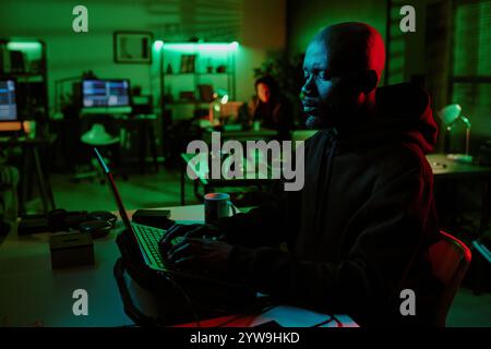 Side view of African American hacker working on laptop with cooling pad in office lit by green lights Stock Photo