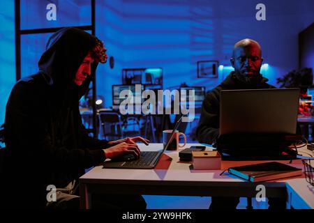 Medium close up of biracial hacker coworkers sitting at table and working on laptops in room illumed with neon lights Stock Photo