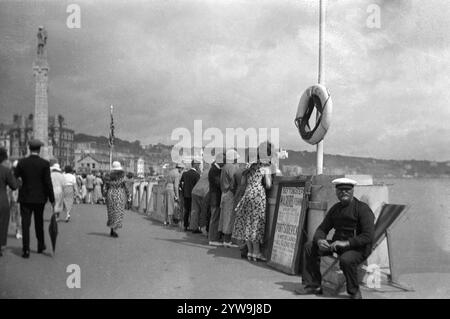 1930s, historical, people at the seafront  Douglas, Isle of Man, England, UK. Fisherman in cap sittng in a deckchair by a sign saying, Stefton Steps, mackerel fishing and day trips to Port Soderick with 'All Glens Free'. Port Soderick beach is a peaceful and tranquil area a few miles from Douglas. Stock Photo