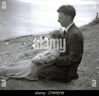 1950s, historical, a young woman in a long skirt & top lying in the lap of a young man wearing a pin-stripe suit, shirt & tie kneeling down on the grassy slope of a hillside at the coast, England, UK. Stock Photo