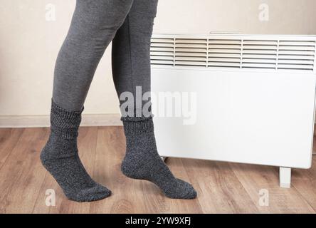 Young woman in warm socks near electric heater at home. Concept of heating season Stock Photo