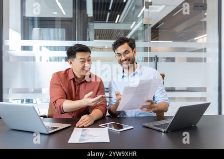 Two colleagues are sitting in a contemporary office discussing paperwork. They are using laptops and a tablet, reflecting a productive and collaborative work environment. Stock Photo