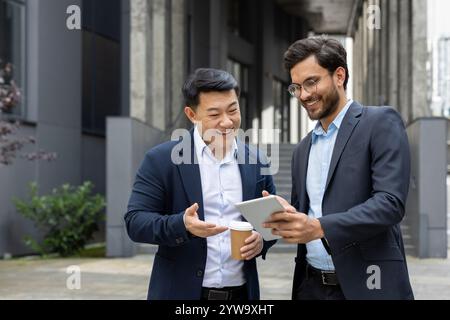Two professional men in suits having a discussion over a tablet outdoors. One is holding a coffee cup, and both are smiling, signifying a positive and collaborative business interaction. Stock Photo