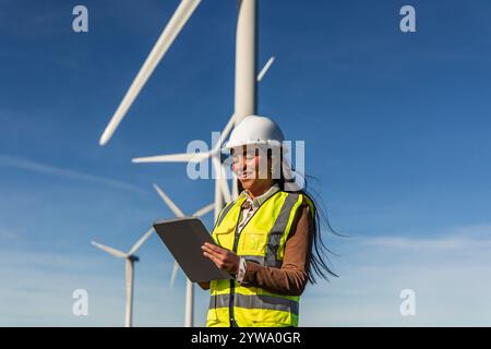 Smiling latin american female engineer wearing safety vest and helmet using digital tablet while working in wind turbine power plant, generating clean Stock Photo