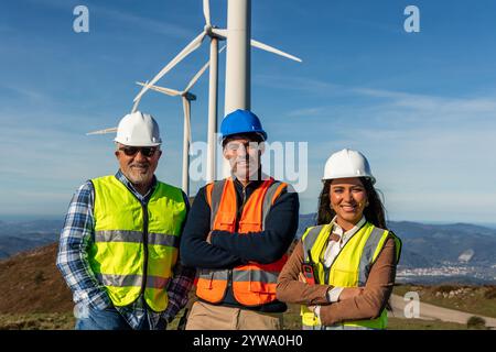 Three multiethnic engineers are wearing safety vests and helmets, standing with their arms crossed in front of wind turbines, representing the growth Stock Photo