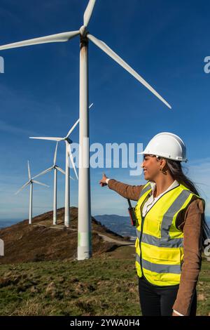 latin Female engineer wearing a safety vest and helmet points towards a group of wind turbines on a grassy hillside, symbolizing the growth and potent Stock Photo