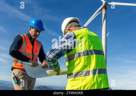 Two engineers wearing safety vests and helmets are reading a blueprint next to a wind turbine, working on a project related to green energy and renewa Stock Photo