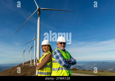 Two multicultural engineers with hardhats and safety vests are posing with arms crossed in front of a group of wind turbines on a mountaintop, represe Stock Photo
